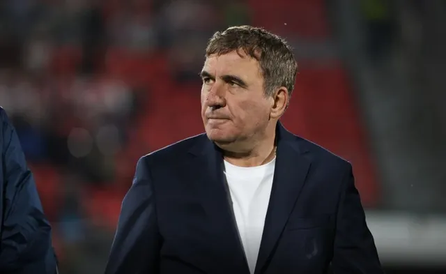 Un bărbat cu părul scurt și cămășă albă poartă un sacou închis la culoare în fața unui fundal cu scaune roșii difuze.