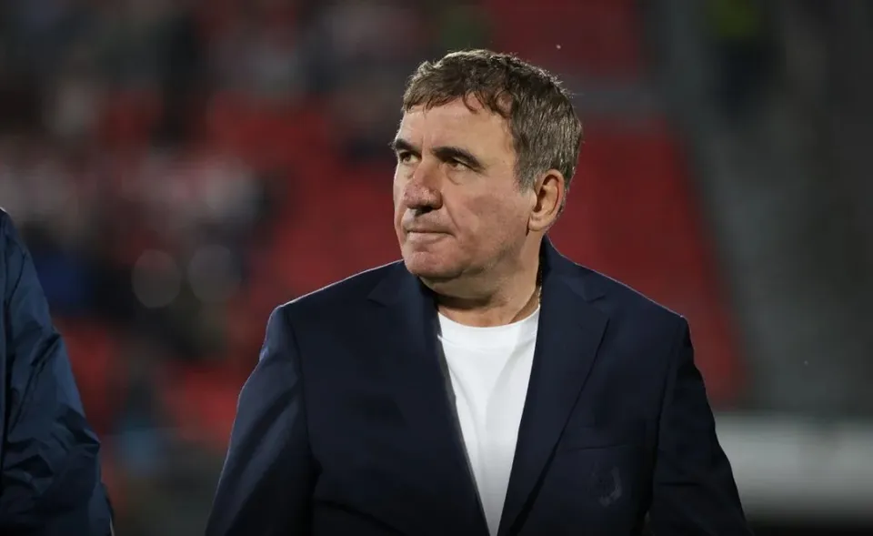 Un bărbat cu părul scurt și cămășă albă poartă un sacou închis la culoare în fața unui fundal cu scaune roșii difuze.