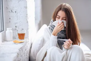 O femeie cu părul lung stă în pat, fiind învelită în pături și ținând un termometru și un șervețel la față.
