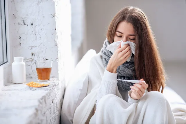 O femeie cu părul lung stă în pat, fiind învelită în pături și ținând un termometru și un șervețel la față.
