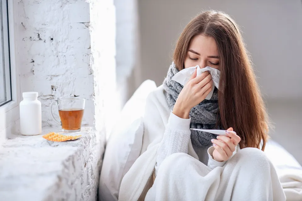 O femeie cu părul lung stă în pat, fiind învelită în pături și ținând un termometru și un șervețel la față.