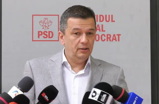 Un bărbat cu părul scurt și sacou gri stă în fața unui panou cu logoul PSD și mai multe microfoane.