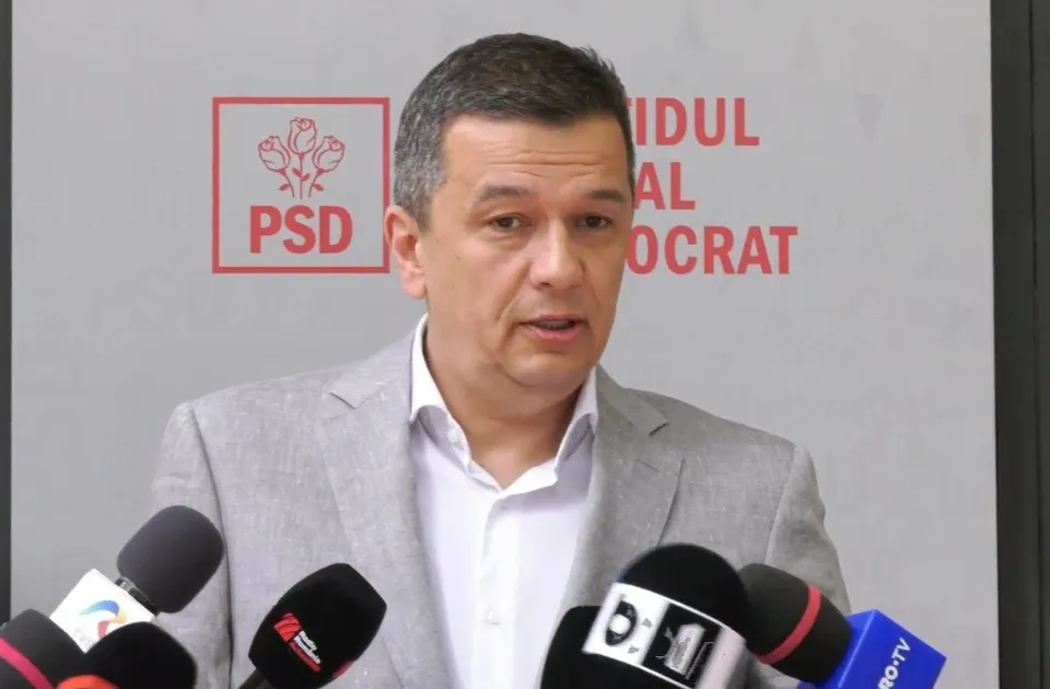 Un bărbat cu părul scurt și sacou gri stă în fața unui panou cu logoul PSD și mai multe microfoane.