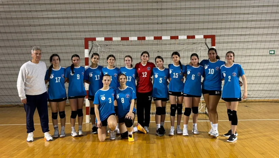 Un grup de femei îmbrăcate în echipamente sportive albastre și roșii stau în linie pe un teren de handbal în fața unui gol.