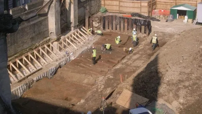 Mai mulți oameni purtând veste reflectorizante lucrează într-o groapă de săpătură de lângă o structură de lemn și un perete de beton.