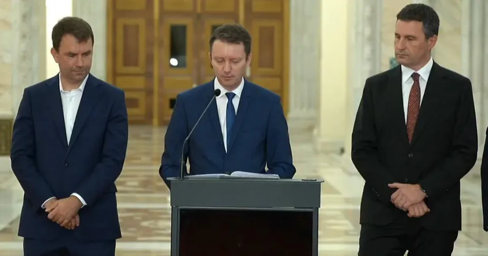Trei bărbați în costume purtând cămași albe stau în picioare lângă un podium gri într-o încăpere cu uși de lemn și coloane albe.
