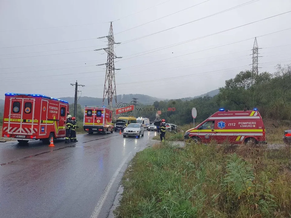 Vehicule de serviciu roșii și albe sunt parcate pe marginea unui drum umed, lângă vegetație, sub un cer înnorat și linii electrice înalte.