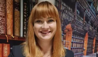 O femeie cu părul roșcat și breton zâmbește în fața unui fundal care înfățișează rafturi de bibliotecă și cărți vechi.