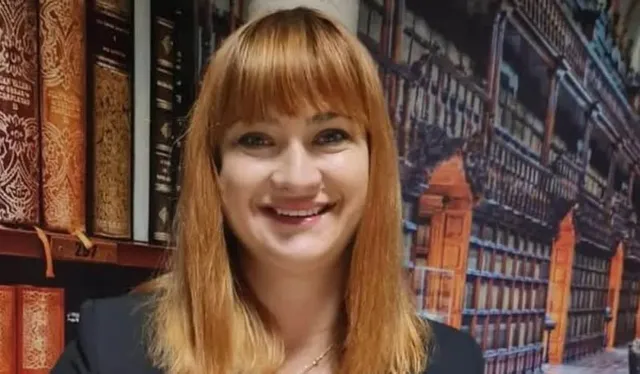 O femeie cu părul roșcat și breton zâmbește în fața unui fundal care înfățișează rafturi de bibliotecă și cărți vechi.