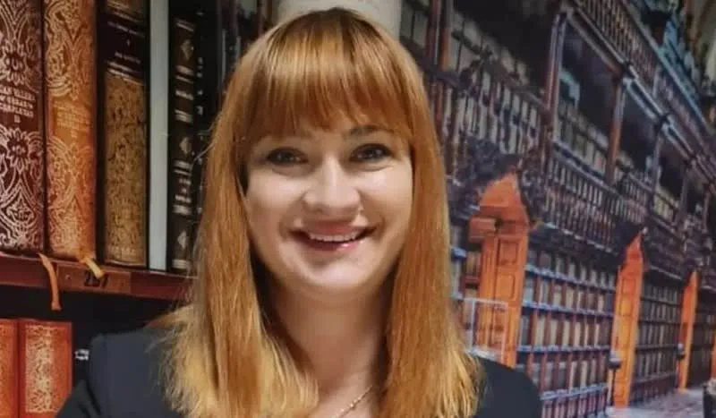 O femeie cu părul roșcat și breton zâmbește în fața unui fundal care înfățișează rafturi de bibliotecă și cărți vechi.