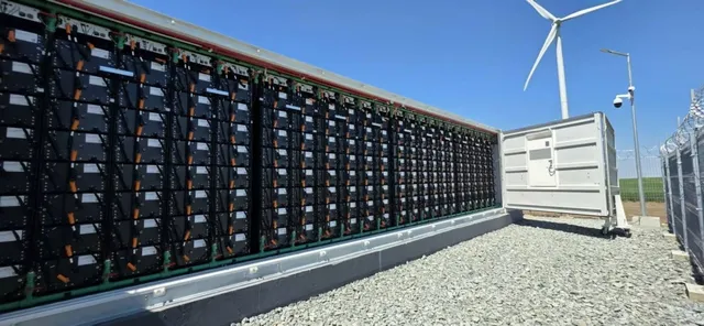 Rânduri de panouri solare sunt instalate pe un suport metalic lângă un container alb și o turbină eoliană sub un cer senin.