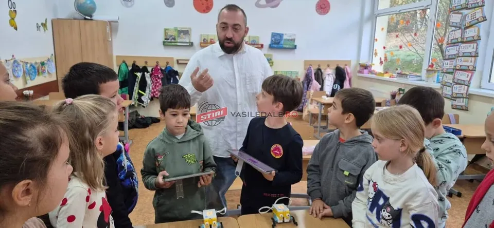 Un bărbat cu barbă stă în picioare lângă un grup de copii care stau în jurul unor mese într-o sală de clasă.
