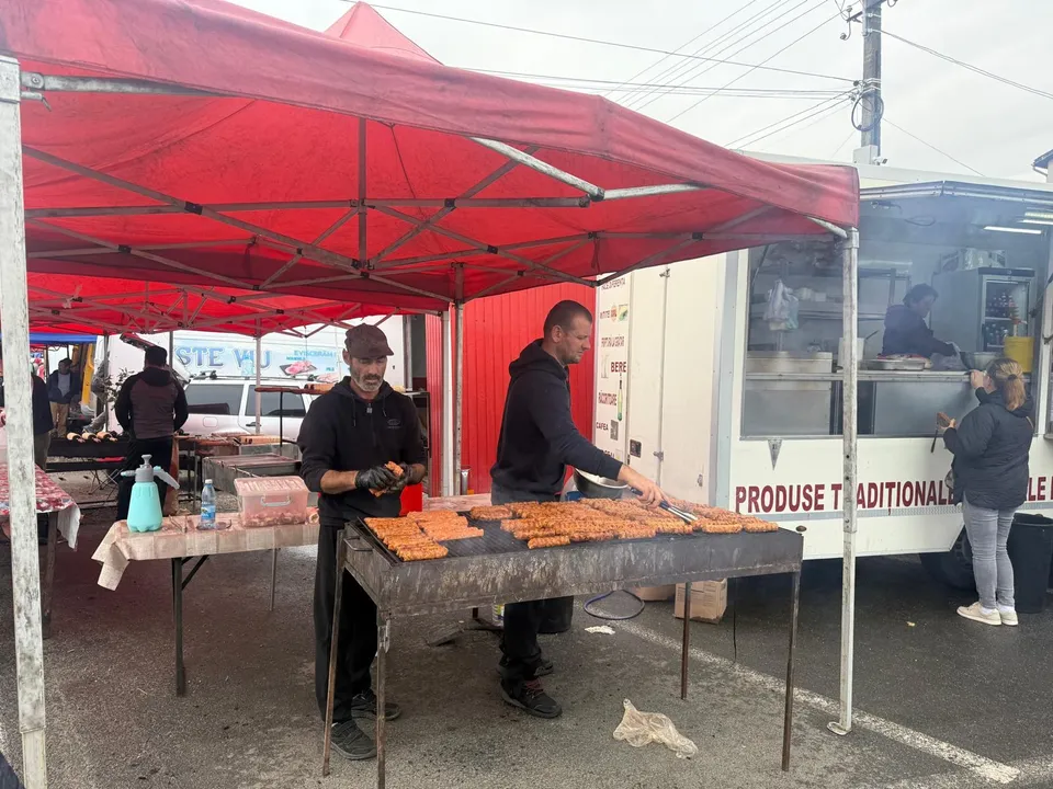 Two men stand under a red canopy preparing food on a large grill, with a white truck displaying text in the background and other market stalls nearby.