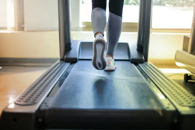 Picioarele unei persoane în pantofi sport se mișcă pe banda de rulare a unui aparat de alergat într-o încăpere luminată.
