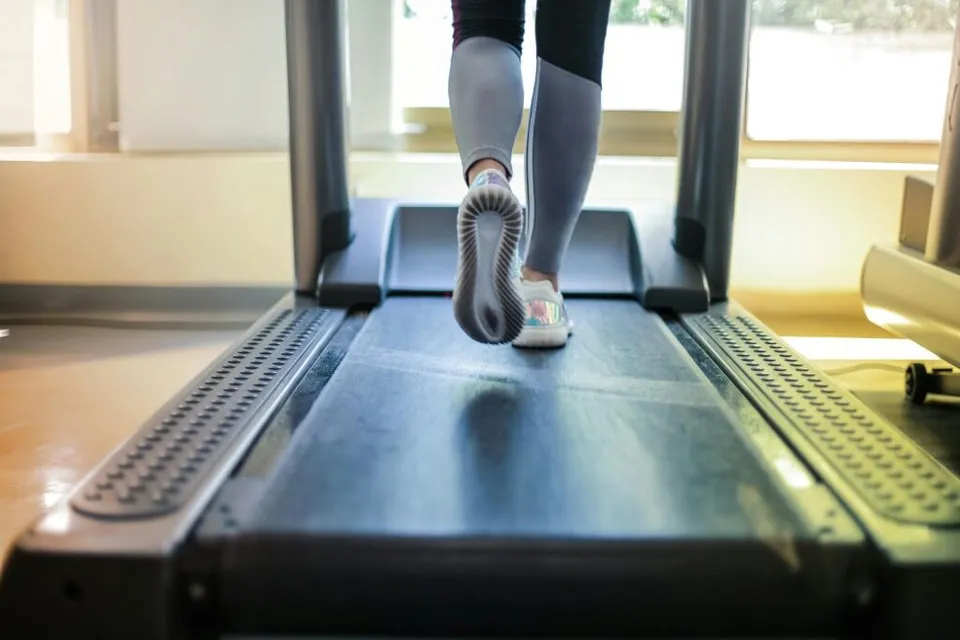 Picioarele unei persoane în pantofi sport se mișcă pe banda de rulare a unui aparat de alergat într-o încăpere luminată.