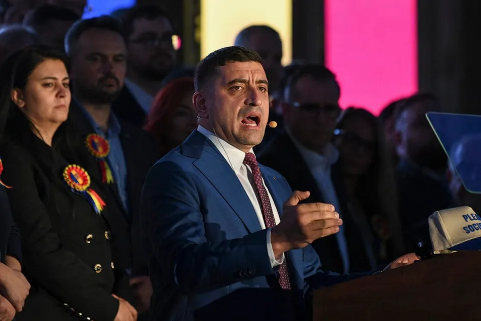 Un bărbat într-un costum albastru și cravată roșie vorbește la un podium, în fața unui grup de persoane.