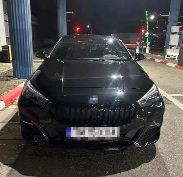 Un autoturism negru este parcat pe un drum asfaltat sub iluminarea nocturnă, având numărul de înmatriculare pixelat.