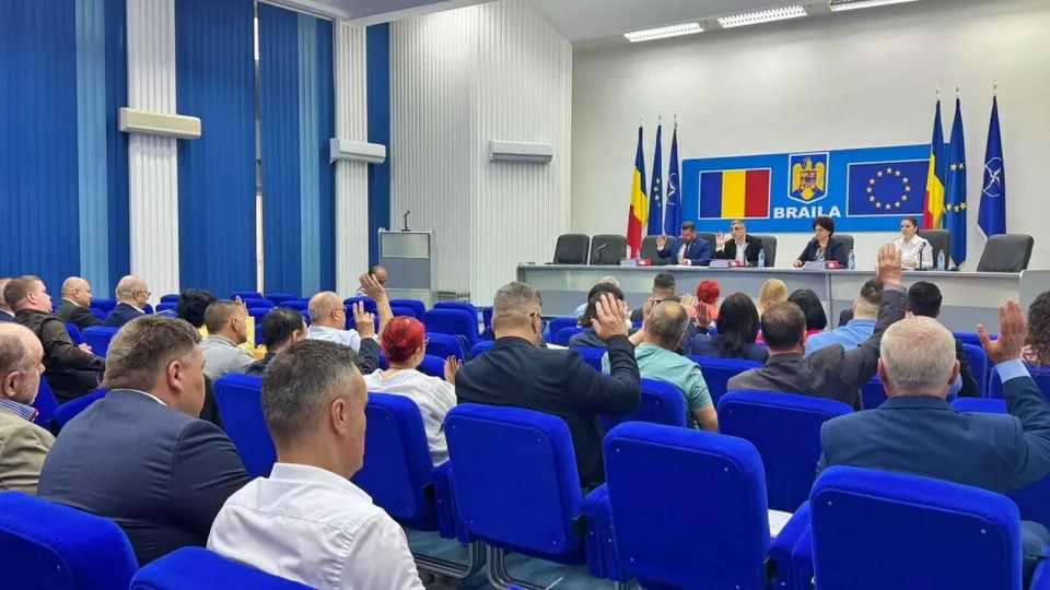 Oamenii stau în scaune albastre într-o sală de ședințe, ridicând mâinile spre o masă lungă unde se află mai mulți oameni și steaguri.