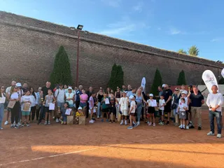 Un grup mare de copii și adulți stă în rând pe un teren de tenis cu nisip, în fața unui zid înalt de cărămidă.