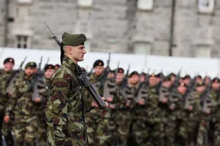 Soldați în uniformă de camuflaj stau în rând, în timp ce un soldat din prim-plan ține un pușcă.