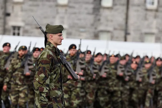 Soldați în uniformă de camuflaj stau în rând, în timp ce un soldat din prim-plan ține un pușcă.
