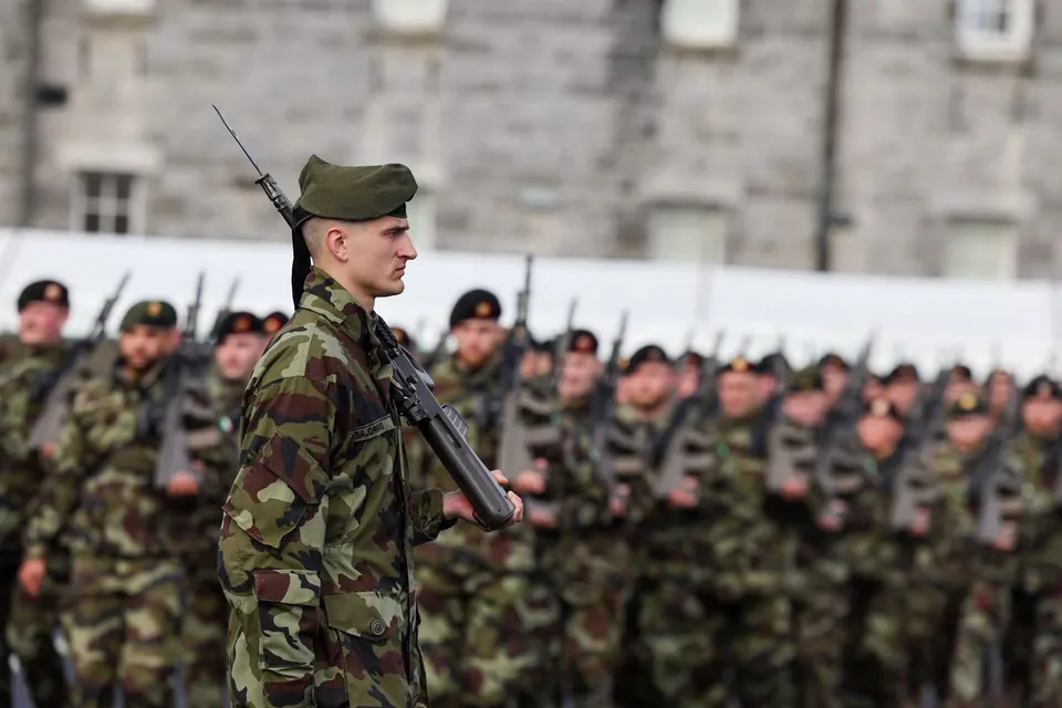 Soldați în uniformă de camuflaj stau în rând, în timp ce un soldat din prim-plan ține un pușcă.