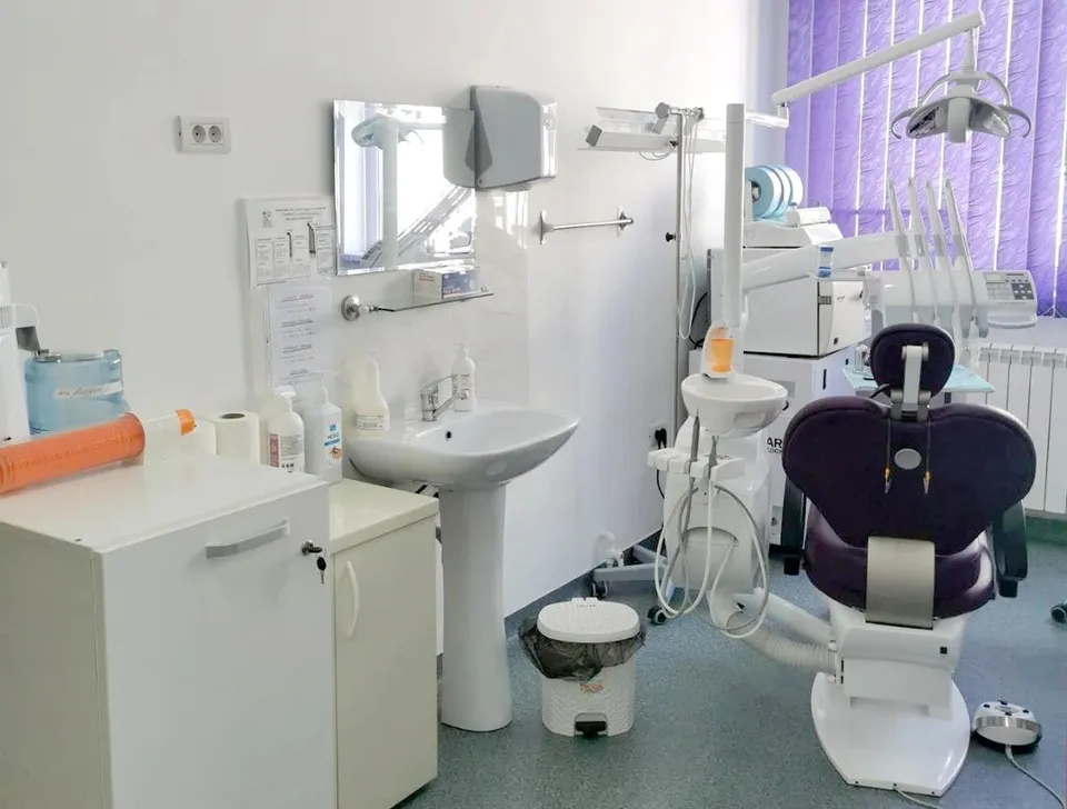 Un cabinet medical conține un scaun de dentist violet, un lavoar alb, un dulap alb și diverse echipamente medicale pe fundal alb.