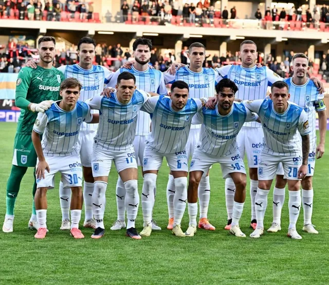 Un grup de fotbaliști în echipamente albastre și verzi stau în rând pe un teren de fotbal verde în fața unor tribune cu spectatori.