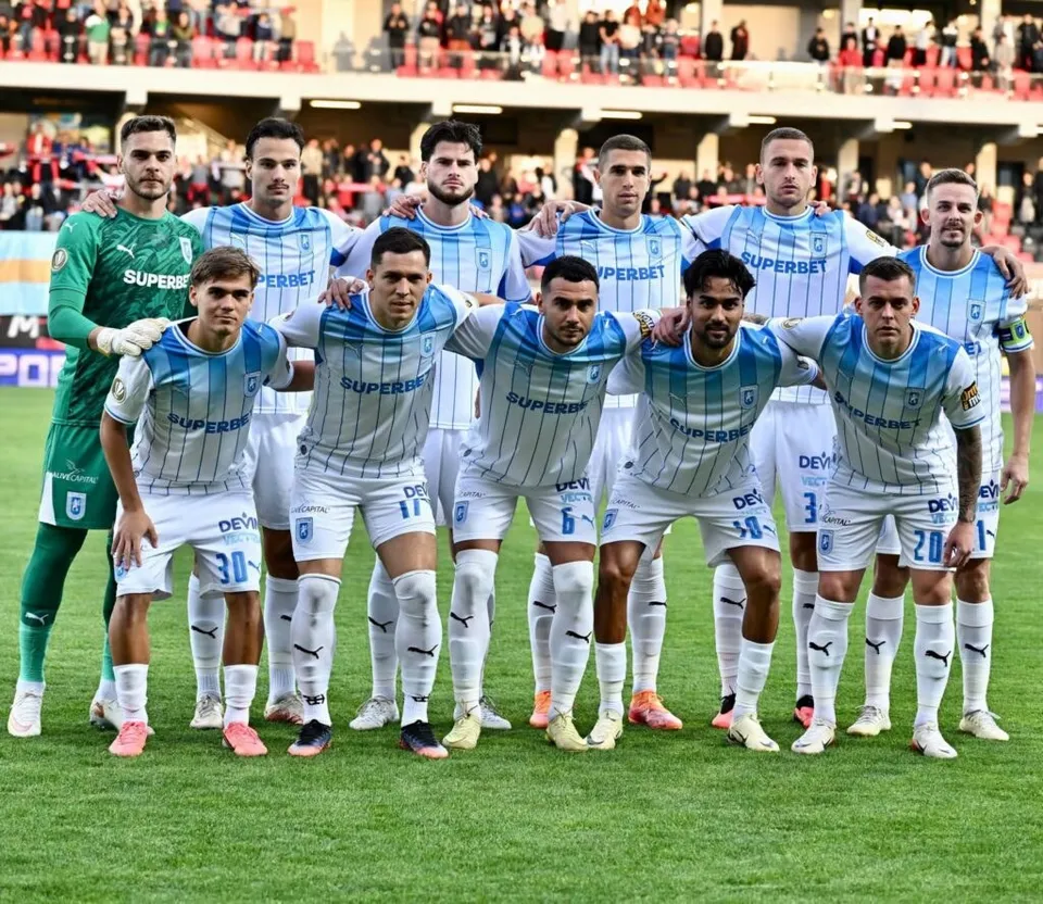 Un grup de fotbaliști în echipamente albastre și verzi stau în rând pe un teren de fotbal verde în fața unor tribune cu spectatori.