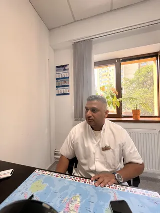 Un bărbat cu cămașă albă stă la o masă lângă o fereastră cu o plantă într-un ghiveci și un calendar pe perete.