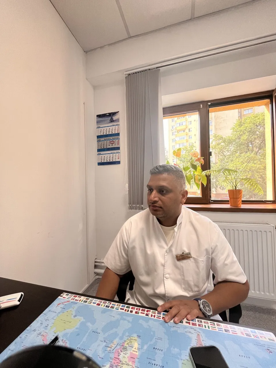 Un bărbat cu cămașă albă stă la o masă lângă o fereastră cu o plantă într-un ghiveci și un calendar pe perete.