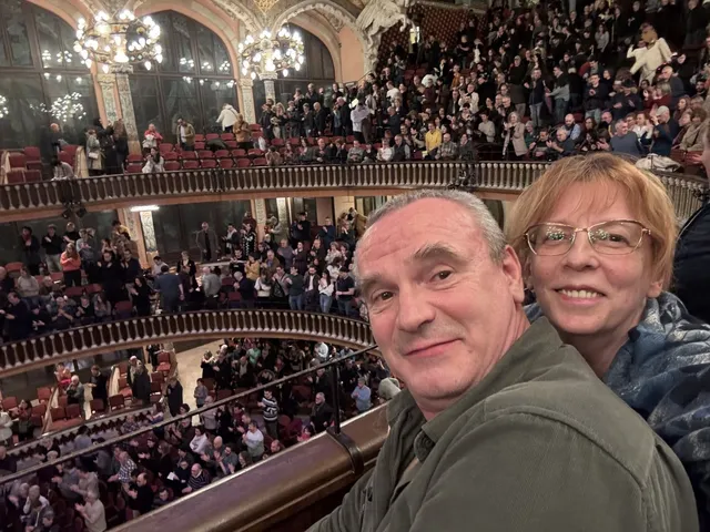 Un bărbat și o femeie cu ochelari zâmbesc în prim-plan, în timp ce o mulțime de oameni ocupă treptele unui auditorium cu balcoane decorate.