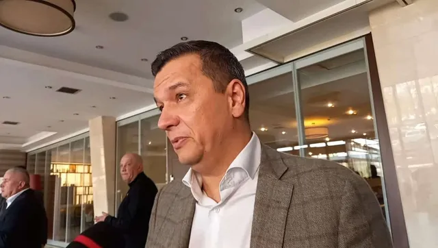 Un bărbat cu părul scurt și sacou gri stă în prim-plan, privind spre stânga, într-un spațiu cu ferestre mari de sticlă.