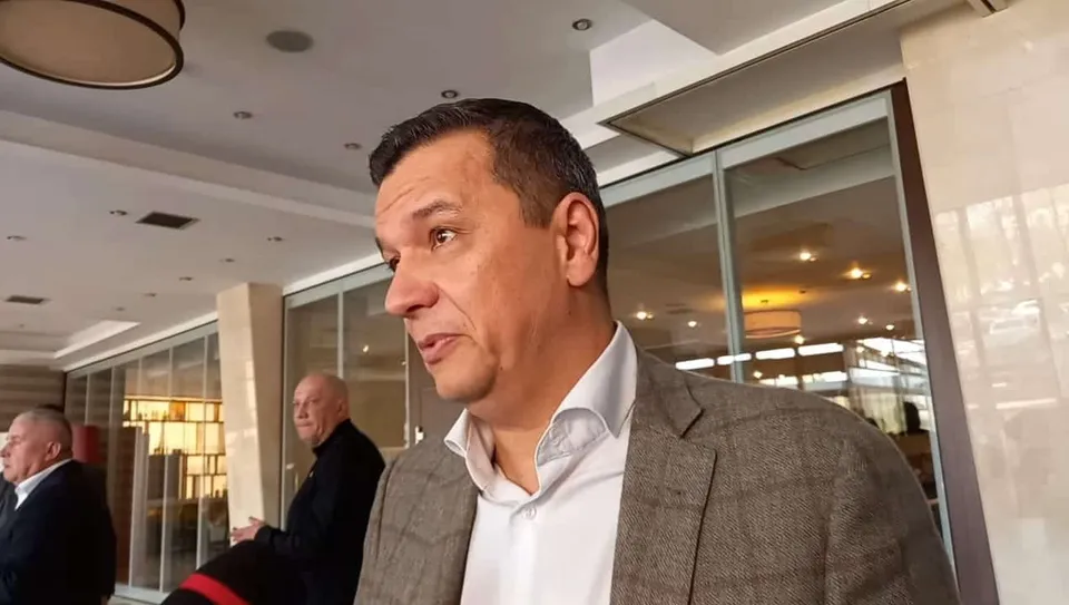 Un bărbat cu părul scurt și sacou gri stă în prim-plan, privind spre stânga, într-un spațiu cu ferestre mari de sticlă.