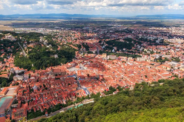 Vedere aeriană asupra unui oraș cu numeroase clădiri cu acoperișuri roșii și zone verzi întinse sub un cer cu nori.