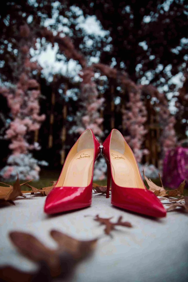 Două pantofi stiletto de culoare roșie sunt așezați pe o suprafață deschisă, având frunze uscate și flori roz în fundal.
