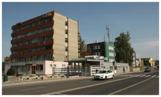 Un clădire înaltă cu fațadă maro și una verde se află lângă un drum asfaltat pe unde circulă o mașină albă.