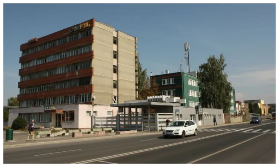 Un clădire înaltă cu fațadă maro și una verde se află lângă un drum asfaltat pe unde circulă o mașină albă.