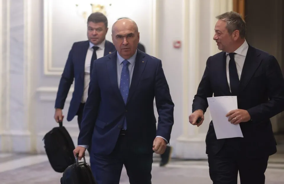 Trei bărbați în costume de culoare închisă merg printr-un hol cu pereți albi, unul purtând o sacoșă și altul un dosar alb.