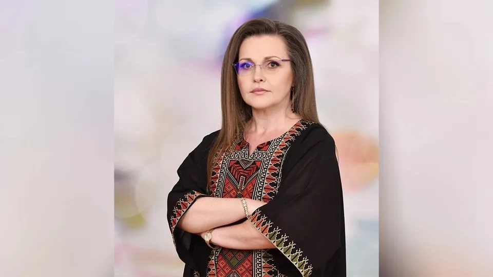O femeie cu ochelari și părul lung, purtând o bluză neagră cu motive colorate, stă cu brațele încrucișate în fața unui fundal difuz.