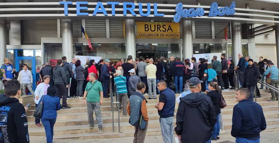 O mulțime de oameni stă în fața unei clădiri cu un afiș care menționează bursa locurilor de muncă.