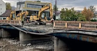 Excavatoare galbene pe un pod sub reparare, cu balustrade din beton și o structură metalică îndoită, pe fundalul unor clădiri și arbori.