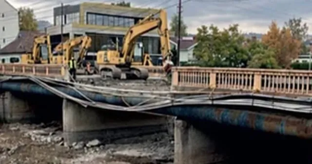 Excavatoare galbene pe un pod sub reparare, cu balustrade din beton și o structură metalică îndoită, pe fundalul unor clădiri și arbori.