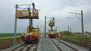 Două platforme mecanice galbene cu operatori lucrează la liniile electrice de deasupra unor căi ferate, în timp ce un lucrător stă pe balast.