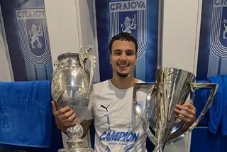 Un bărbat zâmbind poartă un tricou alb și ține două trofee argintii mari în fața unor panouri albastre cu embleme.