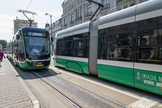 Un tramvai verde și gri circulă pe șine într-o zonă urbană cu clădiri înalte în fundal.