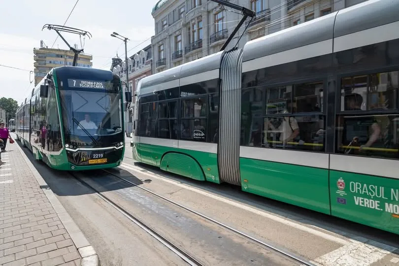 Un tramvai verde și gri circulă pe șine într-o zonă urbană cu clădiri înalte în fundal.