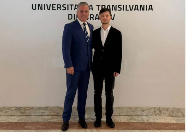 Un bărbat într-un costum albastru și un tânăr într-un costum negru stau alături unul de celălalt în fața unui perete alb cu text negru.