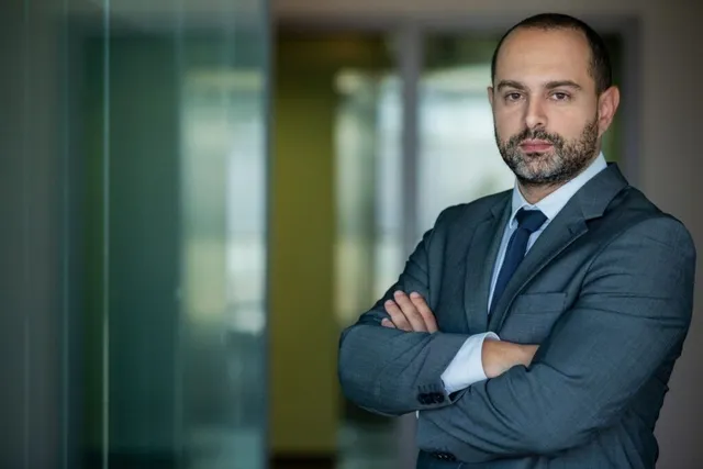 Un bărbat cu barbă poartă un costum gri, cămașă albă și cravată albastră, stând cu brațele încrucișate într-un spațiu de birou.
