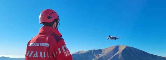 Un bărbat cu cască roșie și jachetă roșie privește un elicopter care zboară deasupra unui munte sub un cer senin.